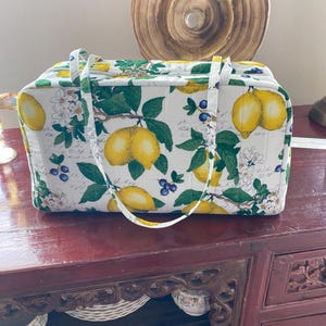 May include: A rectangular quilted fabric bag featuring a white background with a pattern of yellow lemons, green leaves, white blossoms, and small blue berries. The bag has two matching fabric handles and a top zipper closure with metal hardware.