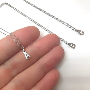 Könnte beinhalten: Halsketten mit silberfarbenen Initialen. Die Halsketten haben feine Ketten mit kleinen Anhängern in Form von Buchstaben. Die Buchstaben A, B und C sind sichtbar. Der Schmuck wird vor weißem Hintergrund präsentiert.