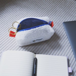 May include: A white fuzzy zippered pouch with a blue and white striped lining. The pouch is open and has a pair of gold scissors inside. The pouch has a red zipper pull and a small white label with the text "Bulle" on it.