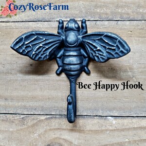 May include: A cast iron bee-shaped hook with a single hook below the bee's body. The hook is dark gray and has the words "Bee Happy Hook" in black text. The background is a light brown wooden surface.