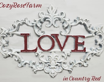 May include: Distressed white wall art featuring the word "LOVE" in red, surrounded by elaborate scrollwork. The text "CozyRoseFarm" is at the top, and "in Country Red" is at the bottom. A decorative piece for the home.