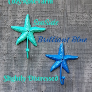 May include: Two starfish-shaped wall hooks in turquoise and blue, with a slightly distressed finish. Each hook has a curved metal base and a round knob at the end. The text "Cozy Rose Farm", "SeaSide", "Brilliant Blue", and "Slightly Distressed" are visible.