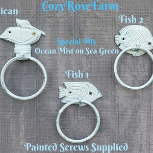 May include: Three cast iron towel rings in a distressed aqua finish. One ring features a pelican design, and the other two are fish-shaped. The text "Cozy Rose Farm" and "Ocean Mist on Sea Green" are visible.