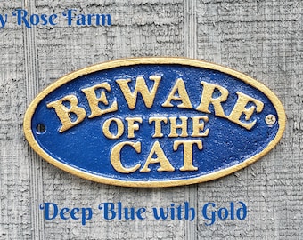 May include: An oval-shaped metal sign with a textured deep blue background and raised gold-coloured lettering that reads BEWARE OF THE CAT. The sign features a gold-coloured border and two screw holes on the sides for mounting purposes.