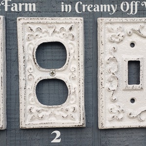 May include: Creamy off-white decorative light switch covers with floral and scrollwork designs. The image shows three different styles: a single switch, a double outlet, and a double switch. The covers are made of metal and have a distressed finish.