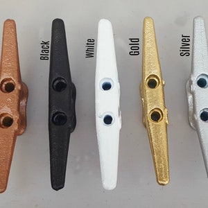 May include: A collection of boat cleats in various metallic finishes: copper, black, white, gold, and silver. Each cleat has a classic design with two holes for securing ropes. The image showcases the different color options available.