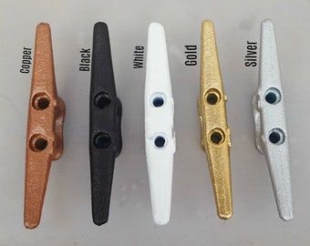 May include: A selection of boat cleats in copper, black, white, gold, and silver finishes. Each cleat features a traditional design with two holes for rope attachment. The image displays the range of colour options available for these marine fittings.
