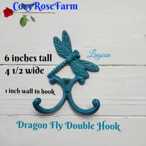 May include: Teal dragonfly double hook, 6 inches tall and 4.5 inches wide, with a 1-inch wall to hook distance. The metal hook features a dragonfly design and the text "Dragon Fly Double Hook". The color is called "Lagoon".