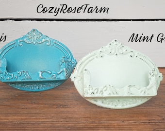 May include: Two ornate, oval-shaped wall shelves in the image. One is a bright turquoise color, labeled "Oasis", and the other is a pale mint green, labeled "Mint Green". Both have a distressed, vintage look and are made of metal. The brand name "CozyRoseFarm" is at the top.