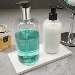 May include: Two bottles of hand wash, one clear with blue liquid and the words "Molton Brown London", the other white. Both are on a white tray. The clear bottle has a silver pump, the white one a black pump.
