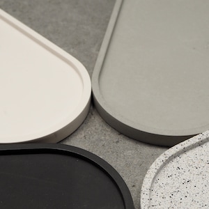 May include: Close-up of four decorative trays in various colours and textures. The trays are oval-shaped with rounded edges. Colours include white, grey, black, and a speckled grey and white. The trays are arranged in a circular pattern.