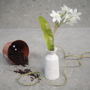 May include: A small white speckled vase holds a sprig of white flowers and a green leaf. A brown plastic flower pot is tipped over, spilling soil. A green twine is draped around the vase.