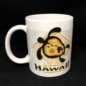 May include: White ceramic coffee mug with a black silhouette of a sea turtle with a map of Hawaii inside the turtle's shell. The mug has a gold design and the word "HAWAII" printed on the bottom.