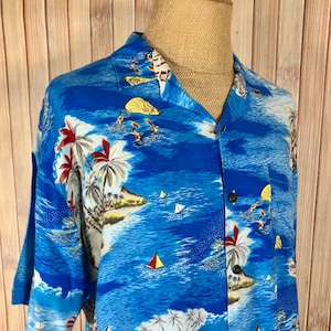 May include: A blue short-sleeved button-down shirt with a tropical print featuring palm trees, sailboats, and surfers. The shirt has a white collar and a pocket on the left side.
