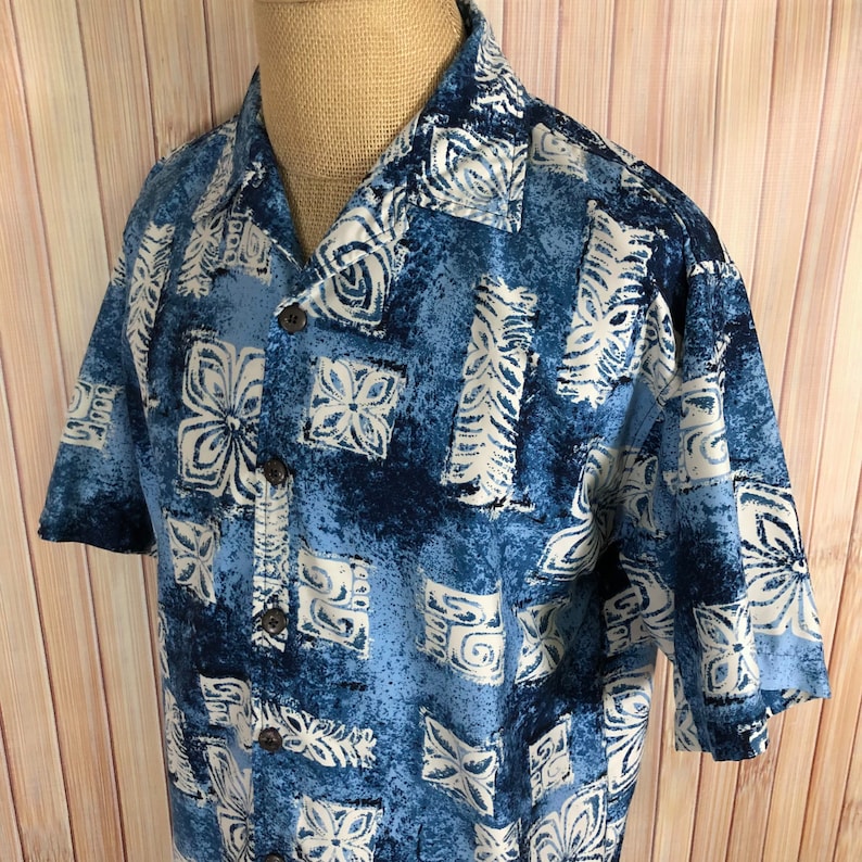 May include: A blue and white patterned short-sleeved button-up shirt with a relaxed fit. The shirt has a tropical print with white flowers and geometric designs on a blue background.