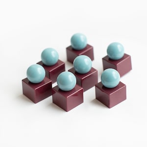May include: A set of seven small decorative objects, each consisting of a smooth, glossy, dark maroon cube base topped with a single, solid, light blue sphere. The pieces are arranged in a cluster, showcasing a uniform design with a polished finish.