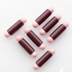 May include: A collection of six cylindrical components, each featuring a dark brown, smooth-surfaced body capped with a small, light pink spherical bead at both ends, arranged in a scattered pattern against a plain white background.