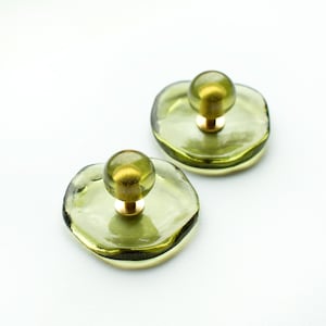 May include: Two olive green glass knobs with a smooth, irregular circular base and a central spherical handle. Each piece features a translucent, light-reflecting surface and a gold-toned metal post connecting the sphere to the base.