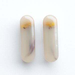 May include: Two elongated, pill-shaped hair clips with a smooth, semi-translucent beige finish. Each clip features a small, pressed yellow flower and a hint of purple petal visible within the resin material, creating a subtle, layered appearance.