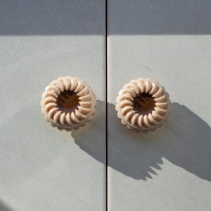 Può includere: Due pomelli rotondi beige per armadi con un design a spirale. I pomelli sono montati su una superficie grigio chiaro, con ombre proiettate sulla superficie. I pomelli hanno un foro centrale di colore più scuro.