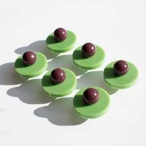 May include: A set of six circular buttons, each featuring a flat, light green disc base topped with a centered, smooth, dark purple spherical bead. The buttons are arranged in a cluster, displaying a consistent design with a glossy finish.