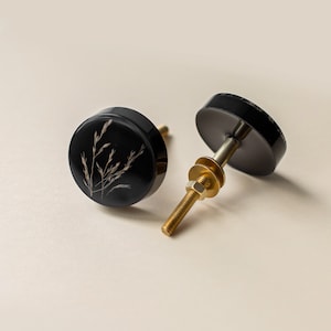 May include: Two black cabinet knobs with a botanical design. Each knob features a round, glossy black surface with a gold-colored stem and hardware. The design on the knob is a delicate, light-colored plant silhouette.