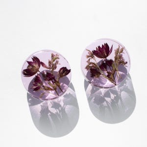 Könnte beinhalten: Zwei runde, lavendelfarbene Harzohrringe. Jeder Ohrring enthält getrocknete, burgunderfarbene Blumen und Stiele. Die Ohrringe werfen einen Schatten auf die weiße Oberfläche darunter.