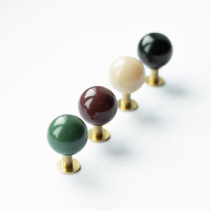 May include: Four decorative cabinet knobs in a row, each with a spherical top. The knobs are in various colors: dark green, burgundy, cream, and black. Each knob has a brass base and stem, suitable for furniture or decor.