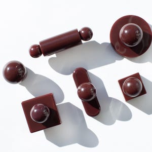Può includere: Una collezione di sei pomelli e maniglie per mobili color marrone scuro lucido in varie forme, tra cui circolari, quadrate, rettangolari e cilindriche. Ogni pezzo presenta un elemento superiore sferico, liscio e arrotondato su una base.