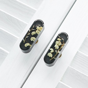 Puede incluir: Dos pomos de armario ovalados negros con un diseño floral. Los pomos presentan flores blancas prensadas en una resina transparente.