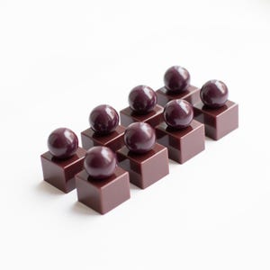 May include: A set of eight dark maroon objects, each consisting of a smooth, glossy sphere securely attached to the top center of a solid, square-based cube. The pieces are arranged in two parallel rows of four, showcasing a uniform, deep red-brown color and a reflective, polished finish on all surfaces.