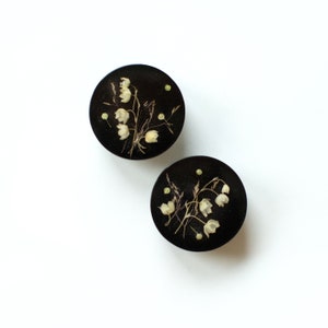 May include: Two black, round decorative objects with a floral design. Each object features a cluster of white flowers and dried grasses. The background is a solid black, and the objects are set against a white background.