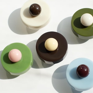 May include: Five circular cabinet knobs with a smooth, glossy finish. Each knob features a flat, round base in various colours including cream, olive green, sage green, dark brown, and light blue, topped with a contrasting spherical centre bead.
