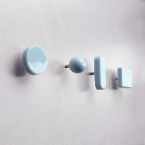 Op de afbeelding: Een set van vier lichtblauwe wandhaken in diverse geometrische vormen, waaronder een grote cirkel, een kleine bol, een verticale rechthoek en een vierkant. Elke haak heeft een gladde, glanzende afwerking en een metalen bevestigingspen aan de achterkant.