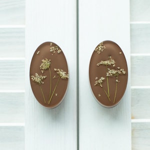 Può includere: Due oggetti decorativi di forma ovale color marrone con fiori bianchi essiccati incorporati. Ogni pezzo presenta una superficie marrone liscia e solida con delicati elementi botanici chiari disposti al centro delle forme ovali.