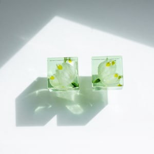 Könnte beinhalten: Ein Paar quadratische Harzohrringe mit einer gepressten weißen Blume darin. Die Ohrringe sind hellgrün.