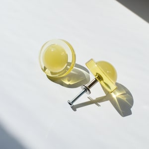 May include: Two round cabinet knobs made of translucent yellow resin. Each knob features a smooth, domed center surrounded by a flat circular rim. One knob is shown from the front, while the other is attached to a silver metal threaded bolt.