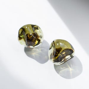 May include: Two rounded, irregular glass objects featuring a translucent olive green hue with deep brown, swirling internal patterns. The glass surfaces are smooth and polished, catching the light to create distinct shadows on the surface below.