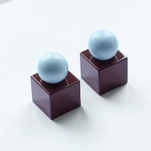 Op de afbeelding: Twee bijpassende oorbellen. Elke oorbel heeft een lichtblauwe bol bovenop een donker bordeauxrode vierkant. De oorbellen staan tegen een witte achtergrond, wat een minimalistische en moderne uitstraling creëert. Het ontwerp is eenvoudig en elegant.
