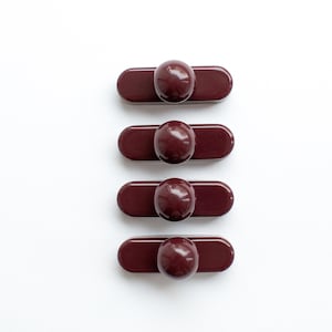 May include: Four dark burgundy buttons, each featuring a smooth, glossy, elongated rectangular base topped with a centred, rounded, dome-shaped protrusion. These uniform, solid-coloured fasteners have a polished finish and a simple, clean design.