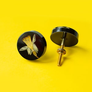 Può includere: Due pomelli rotondi neri per armadio con una vite dorata. Un pomello presenta un motivo di fiore secco pressato.