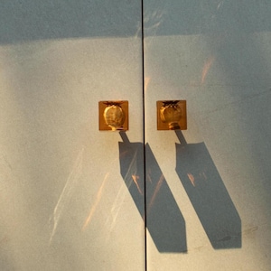 May include: Close-up of a light blue cabinet door with two square, gold-colored knobs. The knobs cast long shadows, and the surface reflects sunlight, creating a play of light and shadow. The cabinet doors are closed.