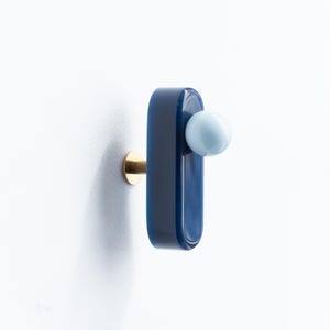 Op de afbeelding: Een moderne wandhaak met een marineblauwe, rechthoekige behuizing en een lichtblauwe bolvormige knop. De haak is gemonteerd op een goudkleurige, cirkelvormige basis. Het ontwerp is minimalistisch en eigentijds.