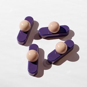 May include: Four individual drawer pulls, each featuring a glossy, deep purple, elongated rectangular base with rounded ends. Centered on each base is a smooth, spherical knob in a soft, pale beige color, creating a clean and modern appearance.