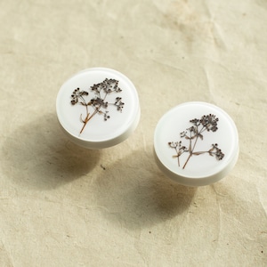 Puede incluir: Dos pomos redondos blancos de resina con flores secas prensadas en su interior. Las flores son marrones y tienen un aspecto delicado y natural.