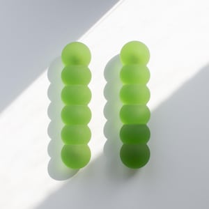 Puede incluir: Dos horquillas verticales, cada una con una fila de seis esferas conectadas de color verde lima esmerilado. Las horquillas tienen una textura mate suave y una apariencia translúcida, creando un diseño lineal y uniforme.