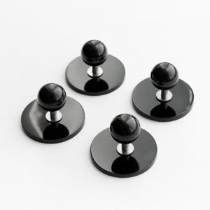 Op de afbeelding: Vier zwarte flessenstoppers met een ronde, glanzende basis en een bolvormige bovenkant. Elke stopper heeft een zilverkleurige steel die de basis en de bovenkant verbindt. De stoppers zijn op een wit oppervlak geplaatst.