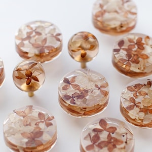 Könnte beinhalten: Sortiment runder, transparenter Harzknöpfe mit eingebetteten Trockenblumen. Die Knöpfe sind in verschiedenen Größen und haben einen Mix aus weißen und braunen Blütenblättern.