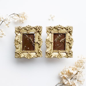 Puede incluir: Dos pequeños marcos de fotos dorados ornamentados con paspartús marrones y flores secas en su interior.