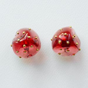 以下が含まれることがあります： 金色のアクセントが付いた2つの赤いガラスビーズ。ビーズは不規則な形をしており、小さな金色の点で覆われています。ビーズは白い背景に配置されています。